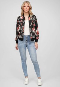 Veste bombardier noire avec un motif floral, manches longues et poignets côtelés. Associée à un jean taille haute bleu clair et des baskets blanches.