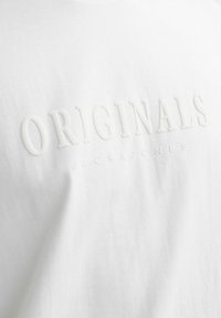 Balta medvilninė marškinėlių su iškilniomis raidėmis, kuriose parašyta "ORIGINALS JACK & JONES" subtilia, derančia spalva. Visiškai lygi tekstūra.