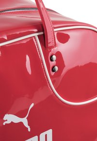 Punainen, kiiltävä duffel-laukku valkoisilla yksityiskohdilla, varustettuna vetoketjulla, ompelutiedoilla ja hopeisella metalliosalla kahvassa. Puma-logo esillä.
