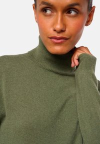 Pull à col roulé vert olive en tissu tricot doux. Comprend un col et des poignets côtelés, avec une coupe ajustée.