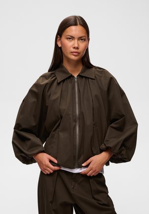 Donna con lunghi capelli scuri che indossa una giacca marrone scuro con cerniera e maniche a sbuffo e pantaloni coordinati su uno sfondo chiaro uniforme.