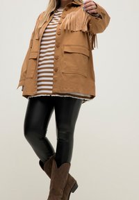 Giacca di pelle scamosciata marrone con bottoni, due tasche frontali, camicia a righe sotto, leggings neri lucidi e stivali da cowboy marroni.