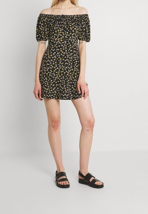 Kobieta ubrana w czarną mini sukienkę z odkrytymi ramionami, bufkami na rękawach i żółto-białym kwiatowym wzorem, zestawioną z czarnymi płaskimi sandałami.