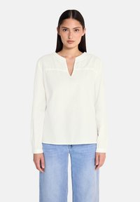 Blusa bianca a maniche lunghe con scollatura a V, caratterizzata da delicate ricami sul petto e maniche con polsini, abbinata a jeans azzurri chiari.