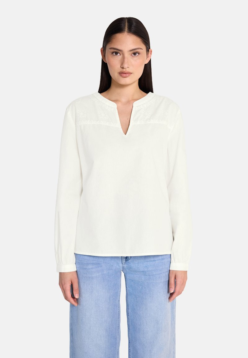 Blusa bianca a maniche lunghe con scollatura a V, caratterizzata da delicate ricami sul petto e maniche con polsini, abbinata a jeans azzurri chiari.