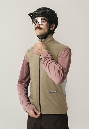 Mann trägt schwarzen Fahrradhelm, Brille, beige ärmellose Weste über rosa langärmligem Shirt, zieht die Weste vor einfachem Hintergrund zu.