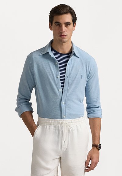 Chemise bleue claire à manches longues en tissu texturé, avec un col et des manches retroussées, superposée sur une chemise rayée bleu marine et blanche. Shorts blancs.
