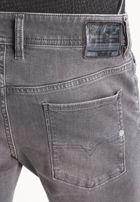 Grå denimjeans med ett läderpatchmärke, detaljerad söm på bakfickan och rak skärning. Slät textur med synliga sömmar.