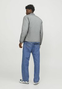 Giacca grigia trapuntata con collo alto e polsini a coste, abbinata a jeans in denim blu e scarpe da ginnastica bianche, vista da dietro.