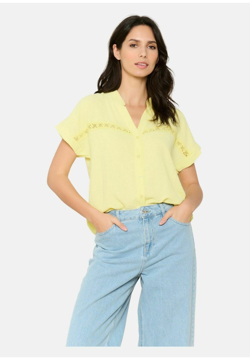 Blusa gialla a maniche corte con chiusura a bottoni e dettagli in pizzo sul petto, abbinata a jeans chiari a vita alta.