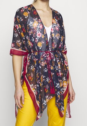 Femme portant un kimono floral transparent avec un bord rouge noué à la taille, par-dessus un haut blanc et un pantalon jaune, visible du cou jusqu’aux hanches.