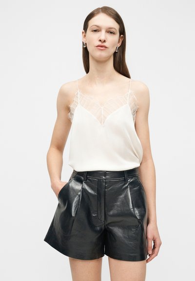 Mladá žena oblečená v bielom tielku bez rukávov s priehľadnými detailmi a čiernych kožených šortkách s vysokým pásom a vreckami, stojaca s jednou rukou v vrecku.