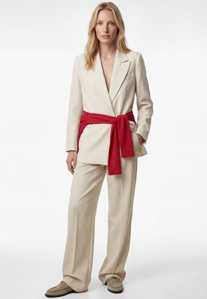 Woman wearing beige suit with wide-leg pants, blazer, red sweater tied around waist, and beige loafers, standing against white background.