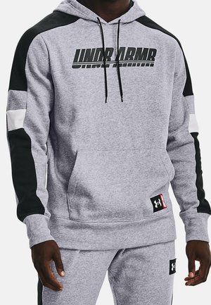Mężczyzna w szarej bluzie z kapturem Under Armour i dopasowanych spodniach dresowych z czarno-białymi panelami na rękawach i przednimi kieszeniami.