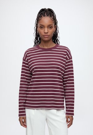 Femme aux cheveux tressés portant un chemisier à manches longues rayé bordeaux et blanc, un pantalon blanc, des chaussures bordeaux et portant un sac à main bordeaux.