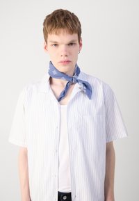 Hvit kortermet skjorte med lyseblå striper og brystlomme, båret over en hvit tanktop. Et blått mønstret bandana er bundet rundt halsen.
