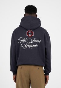 Navy sweatshirt met capuchon, achterkant met een grafisch ontwerp van een rode Zwitserse kruis en witte tekst "The Swiss Yuppie." Zacht materiaal, ontspannen pasvorm.
