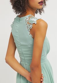 Vihreä mintun värinen mekko, jossa on istuva yläosa, kukkakuvioista pitsiä olkapäillä ja pleated-hame.