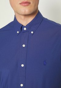 Camicia button-up blu reale con colletto, bottoni bianchi e logo ricamato sul petto. Tessuto dal tocco morbido.