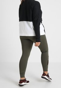 Veste noire courte sur une chemise blanche, associée à des leggings vert olive avec des détails découpés. Chaussures de sport marron avec des accents noirs.