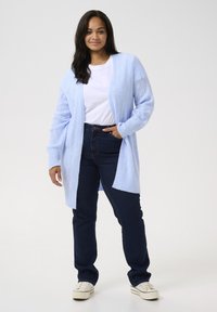 Cardigan en tricot bleu clair avec un motif de feuille, devant ouvert, associé à un jean bleu foncé et des baskets blanches. Le modèle se tient contre un fond uni.
