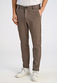 Slanke, bruine broek van zachte stof met een gladde textuur. Voorzien van een knoopsluiting en zijzakken. Gecombineerd met witte sneakers.