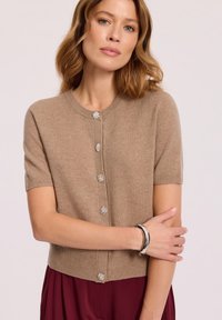 Beżowy kardigan z krótkim rękawem wykonany z miękkiej dzianiny, z metalowymi guzikami w kształcie kwiatów oraz ściągaczem na dole.