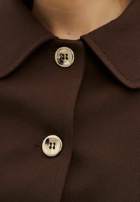 Brun knapp-up skjorta med rundad krage. Har silverfärgade knappar och en mjuk tygtextur. Sömdetaljer längs kragen.