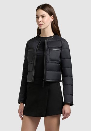 Naine, kes kannab musta tikandiga jope ees taskutega ja musta seelikut, seisab ühevärvilise helehalli tausta ees.