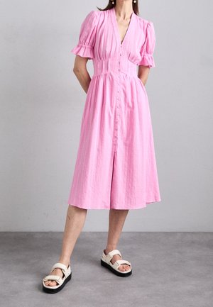 Vrouw staand in een midi-jurk met roze knopen en pofmouwen, met witte platformsandalen tegen een effen achtergrond.