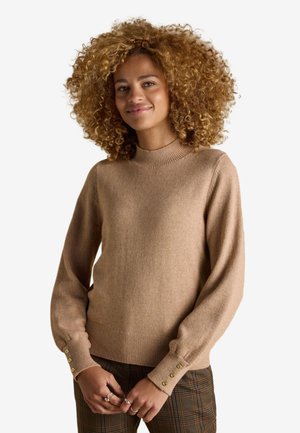 Kvinde med krøllet hår iført en beige langærmet sweater med knapmanchetter og ternede bukser, smilende blidt, hænderne foldet foran.