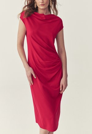 Žena v červenom midi šatách bez rukávov so zdobením na boku a zlatými výraznými náušnicami, stojaca pred jednoduchým svetlým pozadím.