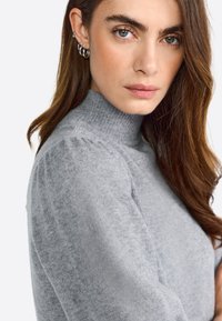 Žena s dugom smešnom kestenjastom kosom koja nosi srebrne kružne naušnice i sivi pleteni roll neck pulover, gleda u kameru.