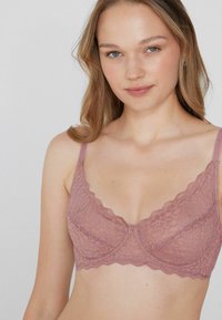 Krajková podprsenka v jemné mauve barvě s ozdobnými vlnkovými okraji. Má tenké ramínka a poloprůhlednou texturu. Jednoduchý design, bez výrazných kovových detailů.