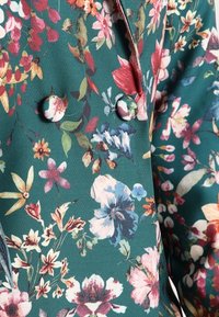 Floral patterned blazer in teal, featuring colorful flowers in red, pink, yellow, and blue. Includes round buttons and a tailored design.