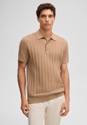 Młody mężczyzna ubrany w beżową prążkowaną koszulkę polo z krótkim rękawem i jasnobeżowe spodnie, stojący na jednolitym szarym tle.