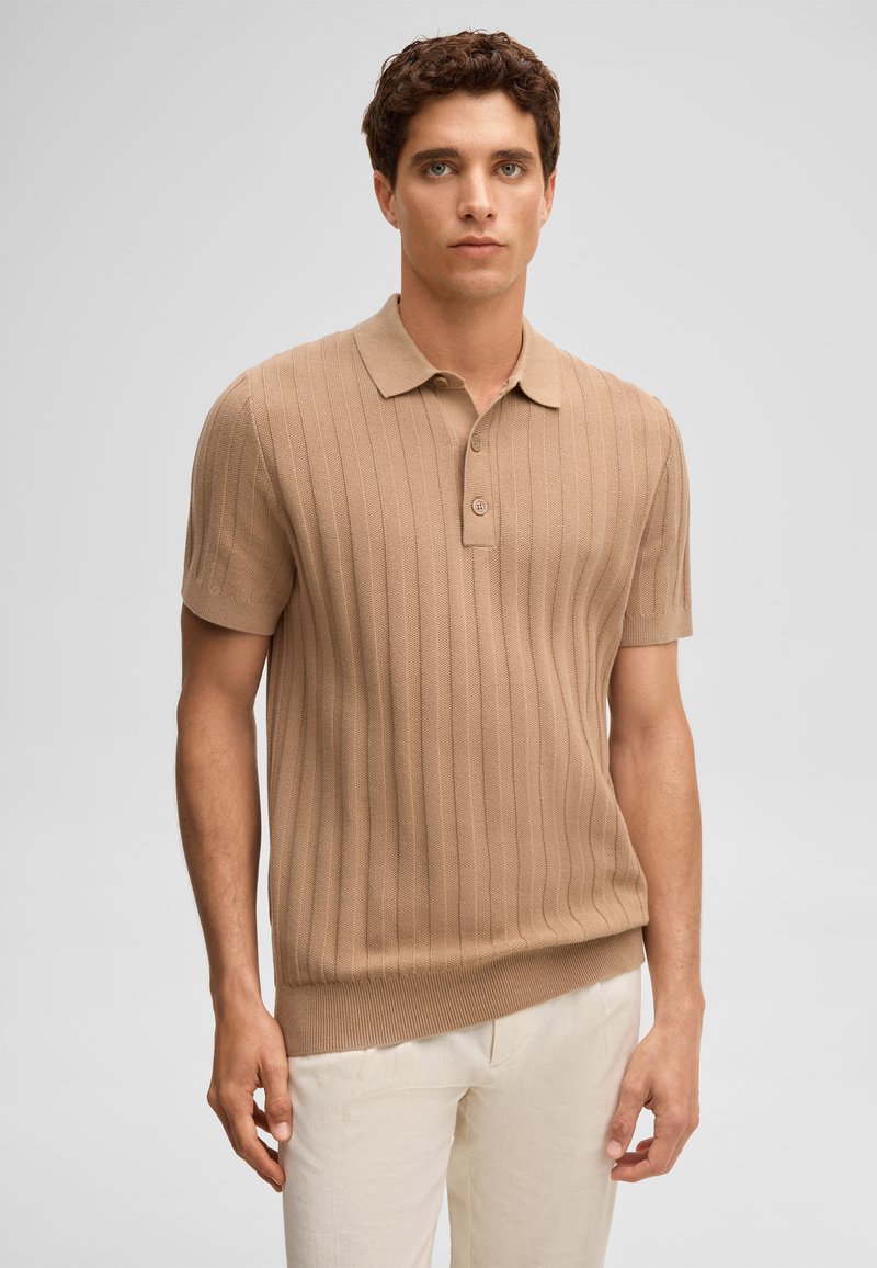 Junger Mann, der ein beige-ripptes Poloshirt mit kurzen Ärmeln und hellbeige Hosen trägt, steht vor einem einfarbigen grauen Hintergrund.