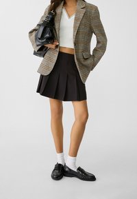 Blazer à carreaux beige et marron, top blanc court, jupe plissée noire, assortis à des mocassins noirs et des chaussettes blanches, tenant un sac noir.