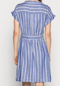 Frau in einem blau-weiß gestreiften Kleid mit passendem Gürtel, von hinten vor einem schlichten hellen Hintergrund gezeigt.