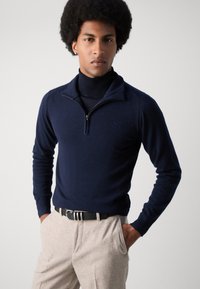 Suéter de marina con cuello con cremallera, hecho de un material suave. Llevado sobre un suéter negro de cuello alto, combinado con pantalones texturizados gris claro.