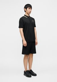 Μαύρο πόλο μπλουζάκι με γιακά που διαθέτει μπεζ λεπτομέρεια και μικρό λογότυπο, συνδυασμένο με μαύρα σορτς από δίχτυ και μαύρα αθλητικά παπούτσια.