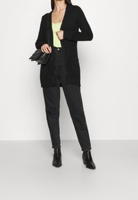 Kvinna iklädd en svart fluffig cardigan, svarta högmidjade jeans, svarta stövlar och en limegrön topp, bärande på en svart kviltad axelväska.