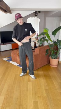 Conjunto casual com uma t-shirt preta com um logótipo, calças largas cinzentas e ténis prateados, situado num espaço interior moderno com plantas.