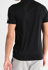 Černé tričko s krátkým rukávem vyrobené z hladkého materiálu s kulatým výstřihem. Na levém rameni se nachází bílé logo.