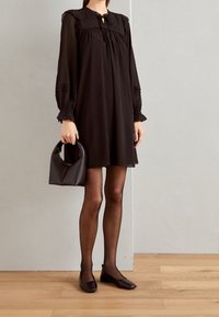 Robe noire, longueur genou avec des manches longues, détails plissés et décolleté en trou de serrure. Associée à un petit sac à main sombre et des chaussures plates.