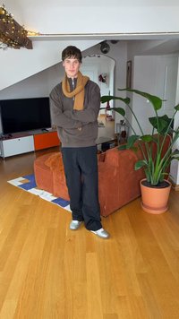 Καφέ φούτερ με κουκούλα, μουσταρδί κασκόλ, μαύρο παντελόνι και ασημένια παπούτσια. Το φόντο περιλαμβάνει πορτοκαλί καναπέ, πράσινο φυτό και ξύλινο πάτωμα.