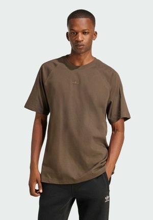 T-shirt in cotone marrone con maniche raglan corte, scollatura rotonda e un piccolo logo tono su tono sul petto. Indossato con pantaloni neri.