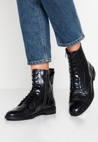 Bottines en cuir noir avec une finition brillante, présentant un laçage à l'avant, une fermeture éclair sur le côté et des détails de bout renforcé. Associées à un denim.
