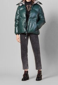 Giacca a vento verde con fodera argentata lucida, dotata di zip frontale, tasche laterali e polsini elasticizzati. Indossata con jeans grigi e stivali neri.
