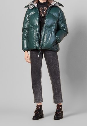 Giacca a vento verde con fodera argentata lucida, dotata di zip frontale, tasche laterali e polsini elasticizzati. Indossata con jeans grigi e stivali neri.
